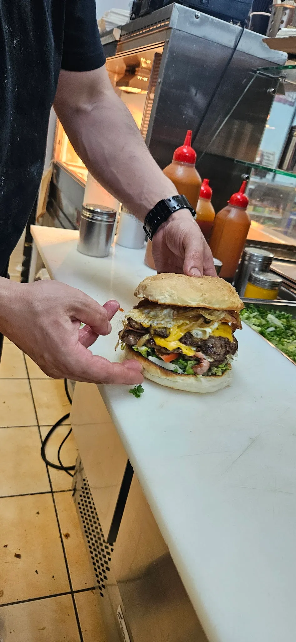 Staff hands presenting a loaded multi-patty burger at the prep counter