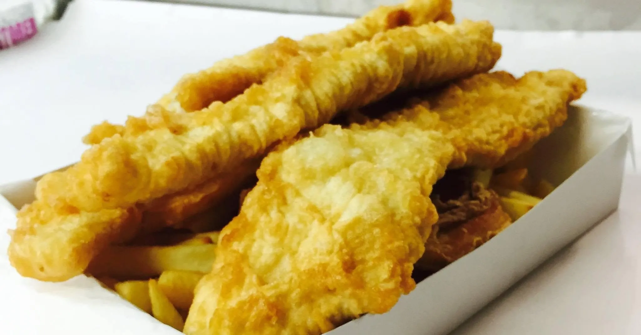 Three golden battered fish fillets over chips in a white takeaway tray